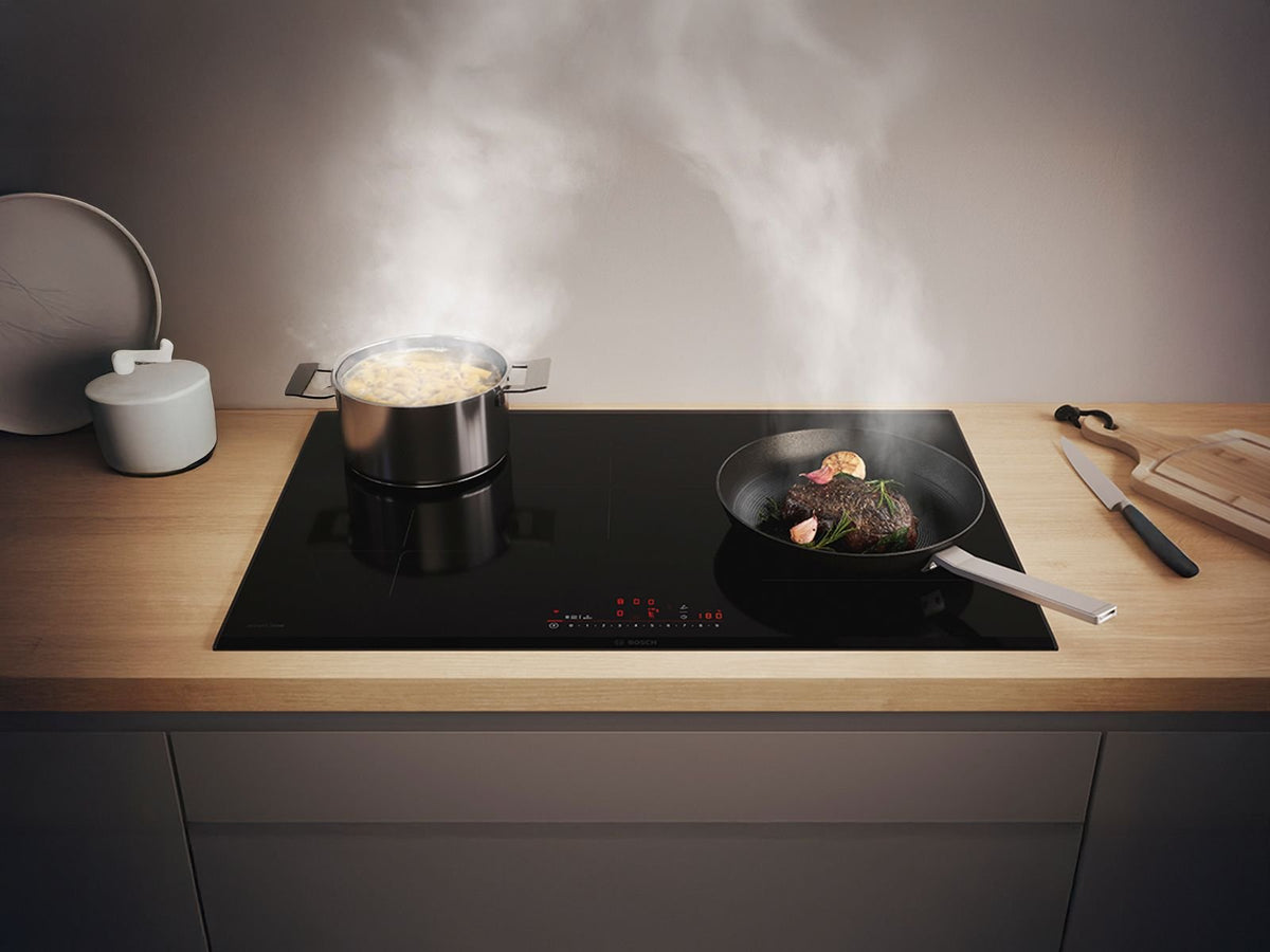 Sort Bosch induktionskogeplade med dampende gryde og pande i en lys bordplade af træ