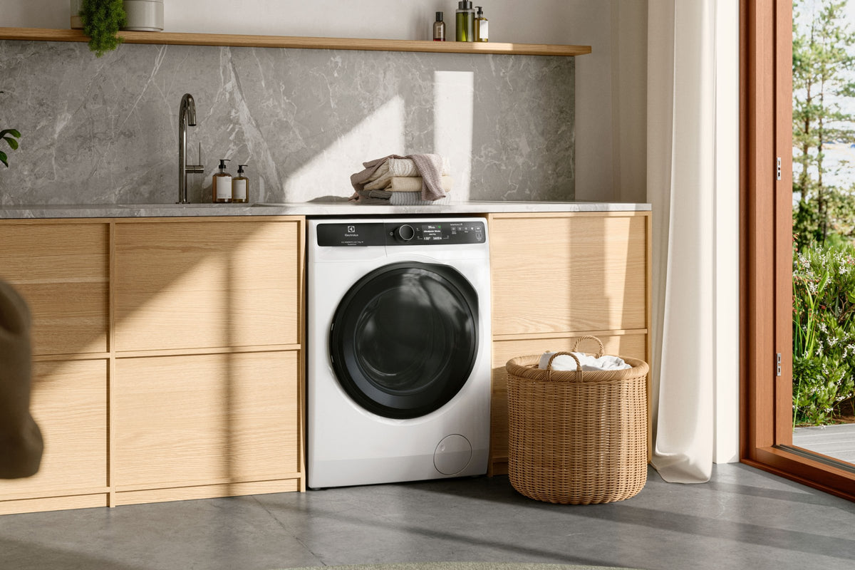 Hvid Electrolux vaskemaskine ved siden af en kurv i et bryggers med store vinduer