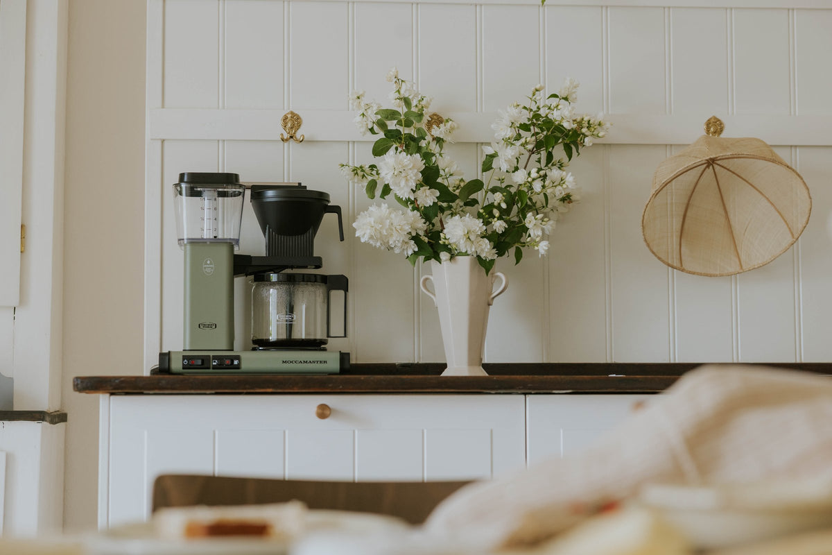 Grøn Moccamaster kaffemaskine på en hvid bordplade med eukalyptusgrene